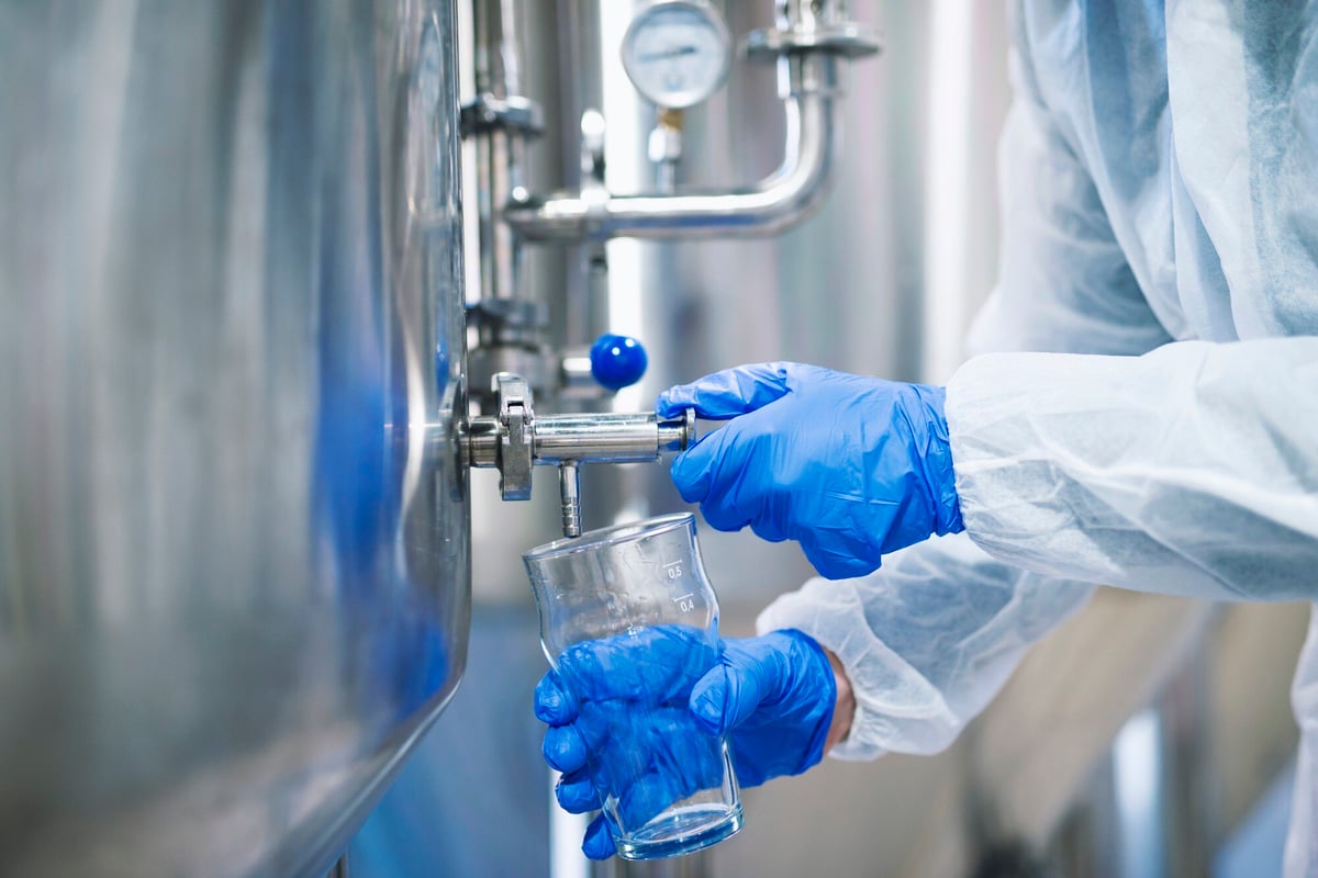 A person in a white protective suit and blue gloves collects a water sample from a stainless steel tank into a glass beaker, demonstrating a step in the water treatment process.